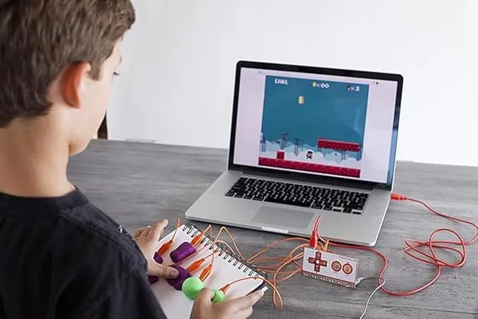 Enfant jouant sur un ordinateur portable en utilisant un kit Makey Makey connecté à des objets en pâte à modeler, posé sur une table.